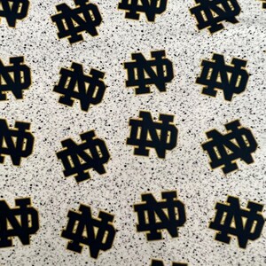 May include: A fabric pattern featuring a repeating dark navy blue Notre Dame monogram logo on a light gray background with a black speckled, granite-like texture throughout the entire surface of the material.