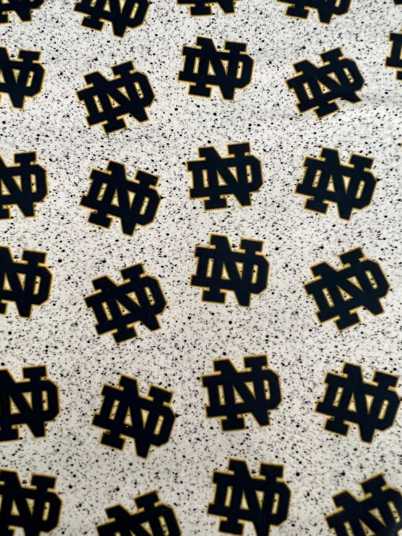 May include: A fabric pattern featuring a repeating dark navy blue Notre Dame monogram logo on a light gray background with a black speckled, granite-like texture throughout the entire surface of the material.