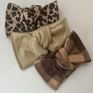 May include: Three knotted headbands in a variety of neutral colors. The top headband features a tan and brown leopard print. The middle headband is a solid tan color. The bottom headband has a brown and tan plaid pattern.