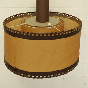 Op de afbeelding: Een bruine en beige hanglamp met een veerkoord en een ronde, gestructureerde kap. De kap heeft een bruine rand met een decoratief stikseldetail.