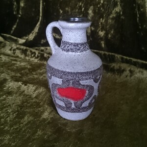 Op de afbeelding: Een witte keramische vaas met een bruin en rood geometrisch ontwerp. De vaas heeft een klein handvat en een rond lichaam.