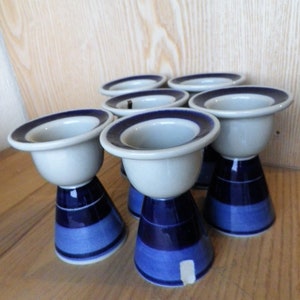Puede incluir: Seis hueveras de cerámica blanca con bases rayadas azules y blancas. Las hueveras están dispuestas en semicírculo sobre una superficie de madera.