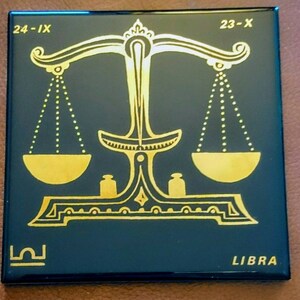 Könnte beinhalten: Eine schwarze quadratische Fliese mit einem goldenen Sternzeichen Waage. Das Symbol ist eine Waage mit zwei Schalen, die an den Armen hängen. Die Fliese zeigt auch die Daten "24-IX" und "23-X" sowie das Wort "LIBRA" in goldenen Buchstaben.