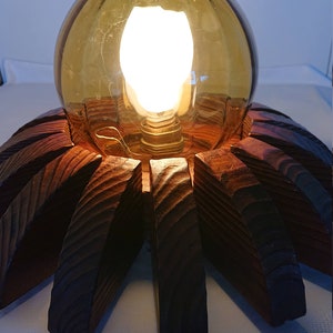 Op de afbeelding: Een houten tafellamp met een bruine, bolvormige glazen kap. De kap heeft een getextureerd oppervlak en een gloeilamp erin. De voet van de lamp is van hout en heeft een stervormig ontwerp.
