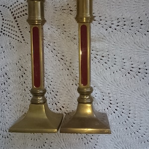 Op de afbeelding: Twee gouden kandelaars met rode accenten. De kandelaars hebben een vierkante basis en een hoog, slank ontwerp.