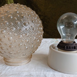 Pode incluir: Um candeeiro de vidro transparente com uma superfície texturizada e com bolhas. O candeeiro está ligado a uma base de cerâmica branca com uma tomada de metal castanho e uma lâmpada transparente.
