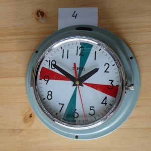 May include: A round, white-faced clock with black hands and red, green, and white markings. The clock has a silver rim and a light blue outer ring. The clock face reads "NKE" and has the numbers 1 through 12.