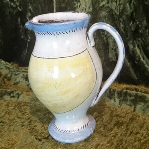 Op de afbeelding: Een witte keramische kan met een geel en blauw ontwerp. De kan heeft een rond lichaam en een uitlopende basis. De handgreep is bevestigd aan de bovenkant van de kan.