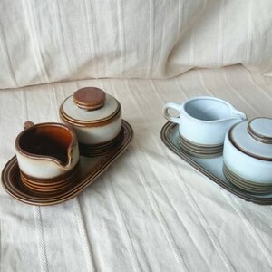 May include: Two sets of ceramic creamer and sugar bowls with brown and white striped designs. Each set is on a matching brown tray.