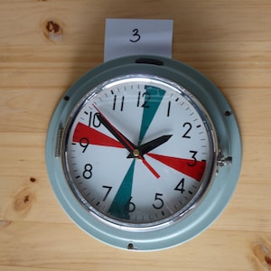 Op de afbeelding: Een vintage, ronde wandklok met een lichtblauwe metalen lijst. De wijzerplaat is wit met zwarte cijfers en wijzers. Rode en teal gekleurde accenten stralen uit het midden. De tijd is ongeveer 9:50 uur.