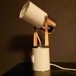 Op de afbeelding: Een witte en koperkleurige spotlamp. De lamp heeft een cilindrische kop en voet, verbonden door koperkleurige steunen. Een schakelaar is zichtbaar op de voet. De lamp is aangesloten op een stopcontact.