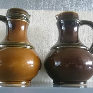 Op de afbeelding: Twee keramische kannen, een bruin en een lichtbruin, met een groene glazuur en een handvat. De kannen hebben een rond lichaam en een smalle hals.