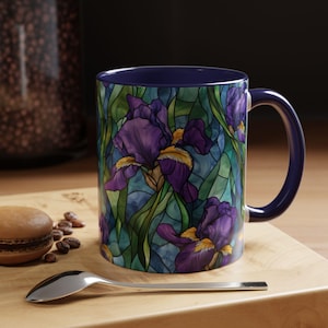 May include: A ceramic mug with a dark blue handle and interior. The mug features a stained-glass style design of purple irises with yellow accents and green leaves against a blue background. The mug sits on a wooden surface.