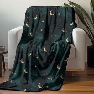 May include: A dark teal throw blanket draped over a white armchair. The blanket features a celestial design with gold crescent moons and stars scattered across the fabric. The blanket's design evokes a night sky theme.