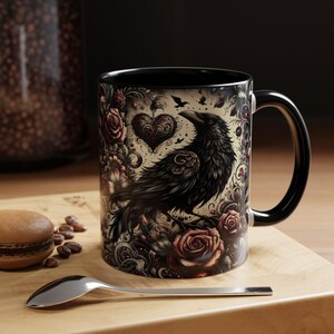 May include: A ceramic mug with a black handle and interior, featuring a detailed illustration of a black raven surrounded by dark red roses, swirling floral patterns, and a decorative heart shape against a cream-colored background.