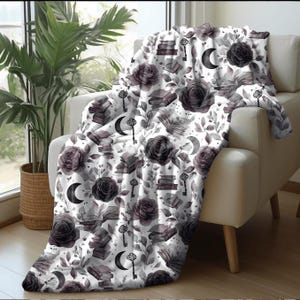 May include: A white throw blanket draped over a cream-colored armchair. The blanket features a repeating pattern of dark gray roses, books, keys, and crescent moons. The blanket is made of a soft material and is suitable for use in a living room or bedroom.