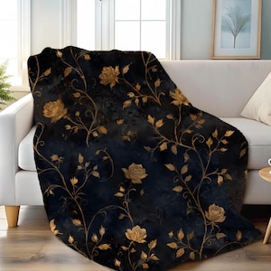 May include: A black throw blanket with a gold floral pattern featuring roses and vines. The blanket is draped over a white sofa, creating a cozy and decorative accent for a living room.
