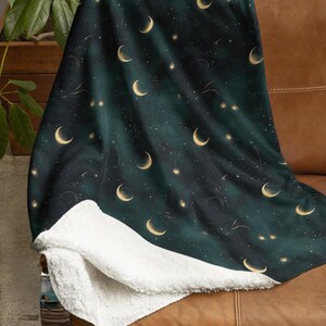 May include: A dark green throw blanket with a celestial design featuring gold crescent moons, stars, and shooting stars. The blanket has a soft, white sherpa lining, and is draped over a brown leather couch.