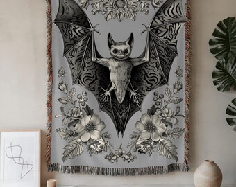Puede incluir: Manta tejida gris con una ilustración detallada de un murciélago con las alas extendidas, rodeado de elementos florales. El diseño del murciélago y las flores es en tonos grises y negros, con un borde con flecos.