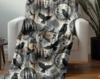 Op de afbeelding: Een decoratieve sprei met een gotisch ontwerp, met zwarte raven, volle manen en boom silhouetten. De sprei is over een witte fauteuil gedrapeerd. Het kleurenpalet omvat grijstinten, zwart en beige, waardoor een spookachtige en sfeervolle esthetiek ontstaat.