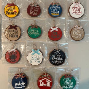 Pode incluir: Uma coleção de enfeites de Natal com vários desenhos e texto. Os enfeites redondos de madeira são decorados com cores festivas e laços. Alguns enfeites apresentam frases como "Santa's Workshop" e "Merry Christmas."