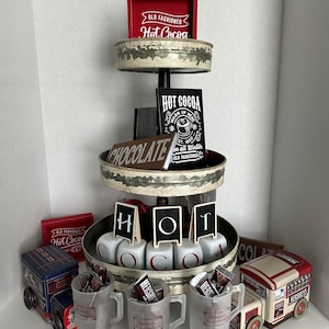 May include: A three-tiered metal stand with a rustic finish, decorated with red and black signs that say "Old Fashioned Hot Cocoa" and "Hot Cocoa". The stand is decorated with chocolate bars, mugs, and other festive items.