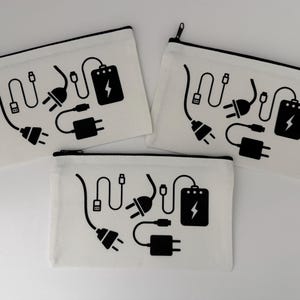 Puede incluir: Tres estuches con cremallera de lona blanca con un gráfico negro de cables de carga, enchufes y un icono de batería. Los estuches tienen una cremallera y un ribete negros.