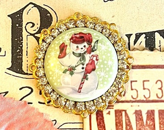 May include: A gold-toned round charm with a clear rhinestone border. The charm features a vintage illustration of a snowman holding a candy cane on a green background with white snowflakes.