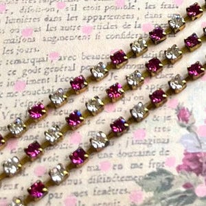 Puede incluir: Primer plano de tres filas de adornos de pedrería. Cada fila alterna entre pedrería transparente y magenta engastada en un metal dorado. El fondo es un papel vintage con texto francés y acentos florales rosas.