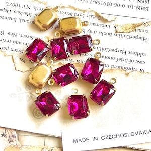 May include: A collection of gold-toned metal jewelry components with rectangular, magenta-colored glass stones. The components are arranged on a vintage paper background, suggesting a craft or jewelry-making theme.