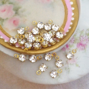 May include: A collection of clear crystal rhinestones with gold-toned jump rings. The rhinestones are scattered on a white surface with a pink and gold design.