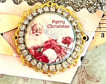 May include: A gold-toned, round, decorative embellishment with a clear, round, glass center featuring a vintage image of Santa Claus with the text "Merry Christmas". The embellishment is surrounded by a ring of clear rhinestones.