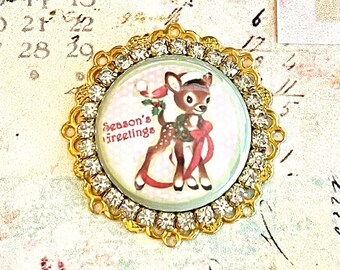 May include: A gold-toned metal button with a white background and a picture of a deer wearing a red Santa hat and a red ribbon. The deer is holding a sprig of holly. The button has a circle of clear rhinestones around the edge. The text "Season's Greetings" is printed on the button.