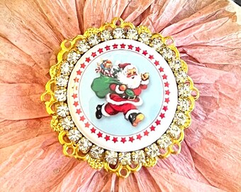 May include: A gold-toned, round, decorative embellishment with a white background and a red border. The center features a vintage illustration of Santa Claus carrying a bag of toys. The embellishment is surrounded by a ring of clear rhinestones.