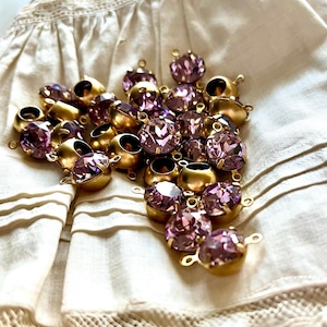 May include: A collection of vintage-style jewellery components. The components feature faceted, light purple crystals set in gold-toned metal settings. Each piece has a small loop for attaching to other jewellery elements. The components are scattered on a cream-coloured fabric.