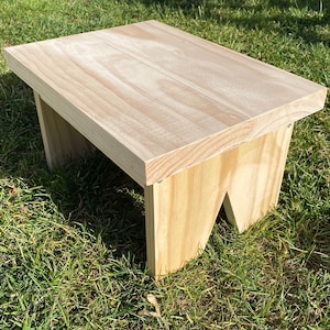 May include: A small, unfinished wooden step stool with a rectangular top and angled legs. The natural wood grain is visible throughout the stool. The stool is sitting on a bed of green grass.