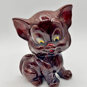 May include: A vintage ceramic cat figurine in a sitting pose. The cat is a glossy, deep burgundy color with yellow eyes and a red nose. The figurine has a classic, cartoonish design.