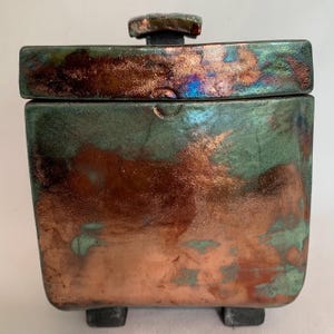 May include: A square ceramic box with a lid, featuring a unique glaze. The box has a copper-colored base that transitions to a teal green near the top. The lid and knob have a similar color scheme. The box sits on four small feet.