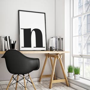 May include: A black and white poster with the letter "n" in a bold font hangs above a wooden desk with a black chair. The desk is supported by two wooden sawhorses. There are two potted plants on the floor next to the desk.