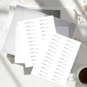May include: White multiplication worksheets with black text, featuring math problems like "8 x 10 = _" and "2 x 3 = _". The worksheets are stacked on a light-colored surface, with a cup of coffee visible in the corner.