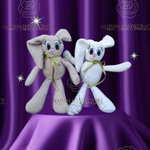 May include: Two plush bunny toys, one beige and one white, with embroidered eyes and pink noses. Both bunnies have long ears and are standing on a purple fabric surface. The text "MADAM HANDYCRAFT" is visible in the background.