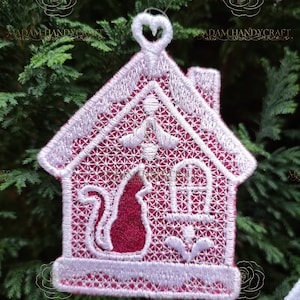 May include: White and pink embroidered Christmas ornament shaped like a house with a cat inside. The ornament has a heart-shaped loop at the top for hanging.