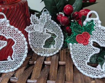 Op de afbeelding: Drie witte kanten kerstversieringen met een konijnen-silhouetontwerp. Een versiering heeft een groen hulstblad en rood bessenontwerp.