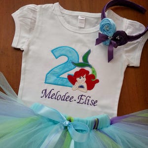 May include: A white short-sleeved t-shirt featuring a blue number two, a mermaid graphic, and the name Melodee-Elise embroidered in purple. The set includes a multi-layered tulle tutu in shades of blue, green, and purple, and a matching headband.