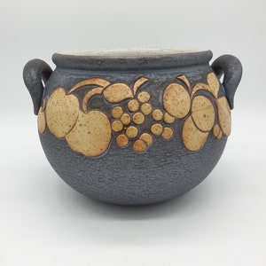 Op de afbeelding: Een grijze keramische pot met een getextureerd oppervlak en een ontwerp van fruit en druiven in bruin en geel.