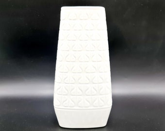 Może przedstawiać: Biały ceramiczny wazon z geometrycznym wzorem. Wazon ma kwadratową podstawę i wysokie, prostokątne ciało.