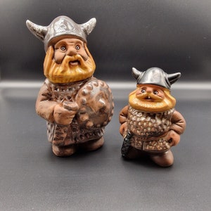 May include: Two ceramic Viking figurines, one larger than the other. The larger figurine is wearing a horned helmet and holding a shield. The smaller figurine is wearing a horned helmet and holding a sword. Both figurines are painted in brown, yellow, and black.