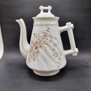 以下が含まれることがあります： 金色の花の模様が施された白い磁器製のティーポット。ティーポットには取っ手と注ぎ口があります。ティーポットは、金色の花と葉の渦巻き模様で飾られています。