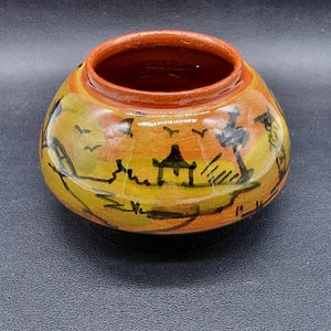 May include: A small, round, orange ceramic pot with a black and yellow painted scene of a pagoda and trees. The pot has a wide opening and a smooth, glazed finish.