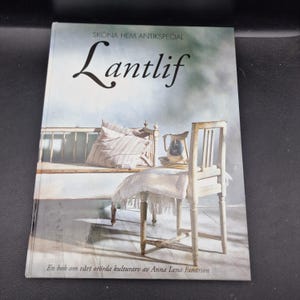 May include: A book titled "Lantlif" with a cover illustration of a rustic interior. The image shows a bed, a chair, and a pitcher. The title is in a decorative font. The book is a special edition.